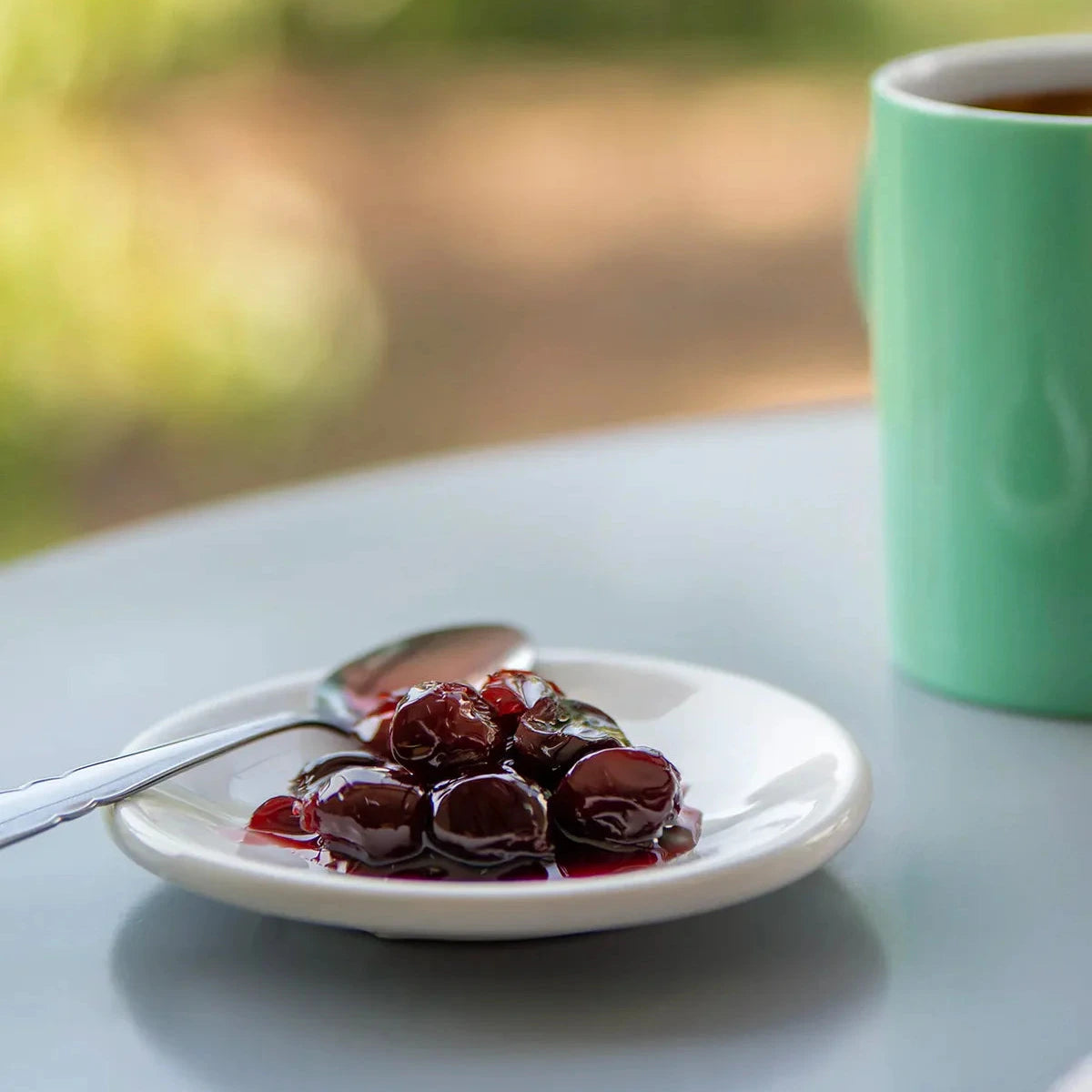Plate of cherries with a spoon on a table next to a green mug.