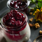 Glass jar filled with a layered dessert topped with red fruit compote on a dark surface.
