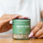 Person holding a jar of sea salt blend with basil and garlic on a wooden surface.