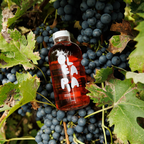 Bottle of red liquid with a white cap on a background of dark grapes and green leaves.
