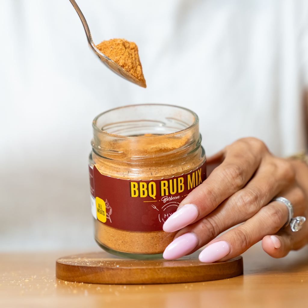 Jar of BBQ Rub Mix with a spoonful being held above, on a wooden surface.