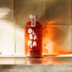 Red bottle labeled 'Olea' on a reflective surface with a tiled wall background