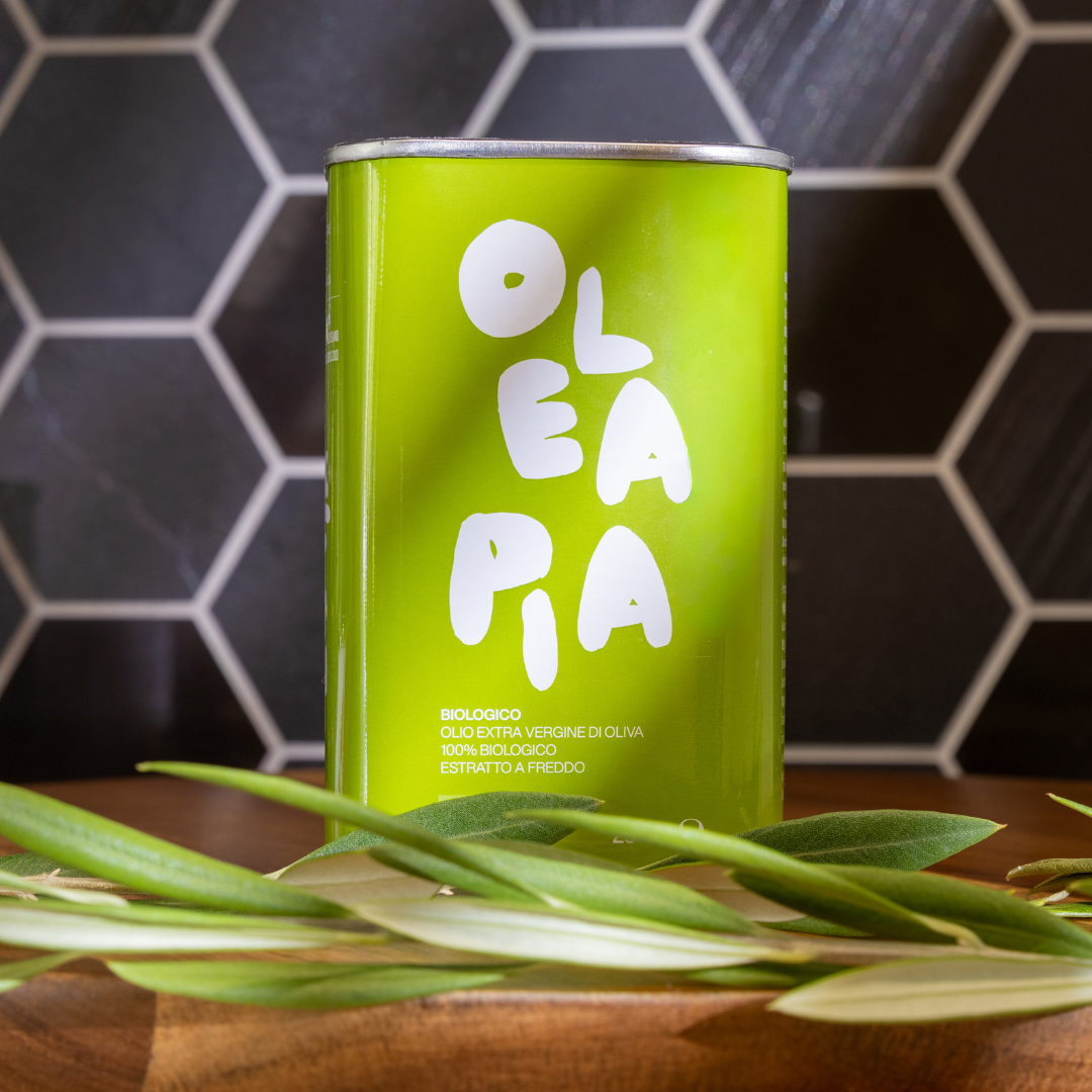 Green canister with 'Olea Pa' branding on a wooden surface with olive leaves, against a hexagonal tile wall.