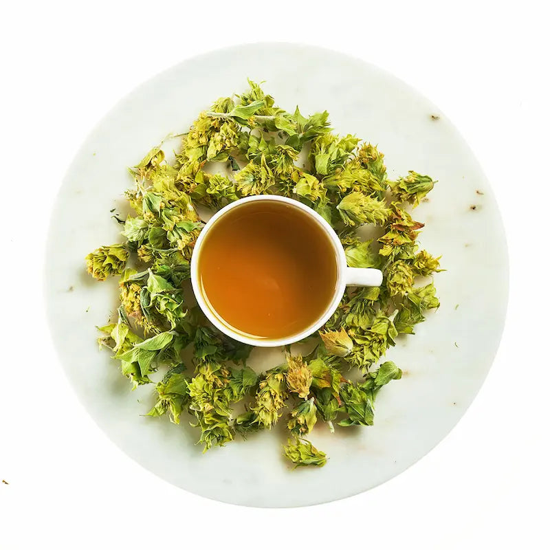 White cup of tea surrounded by green tea leaves on a white plate